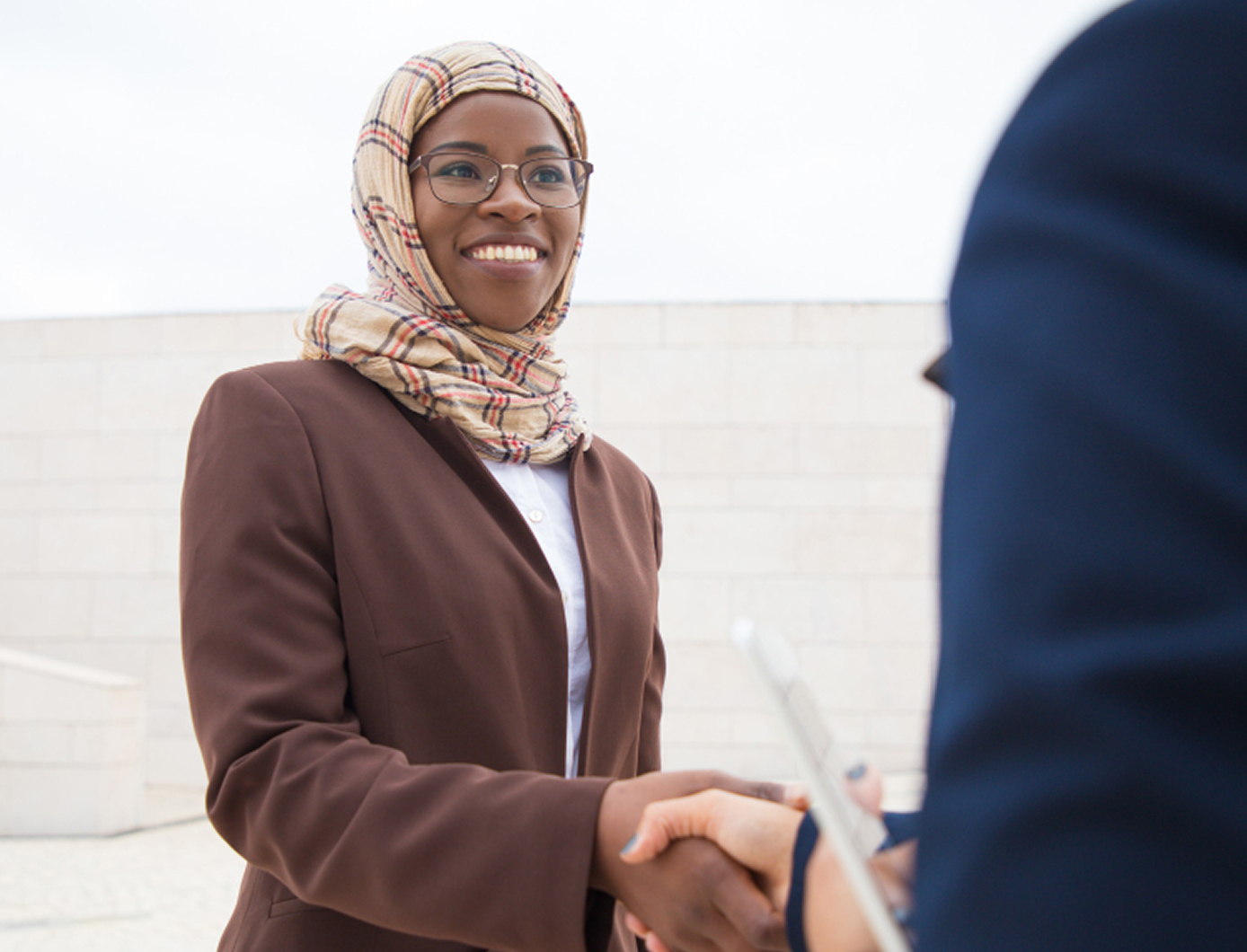 A woman in a hijab and brown blazer shaking hands with a colleague holding a tablet